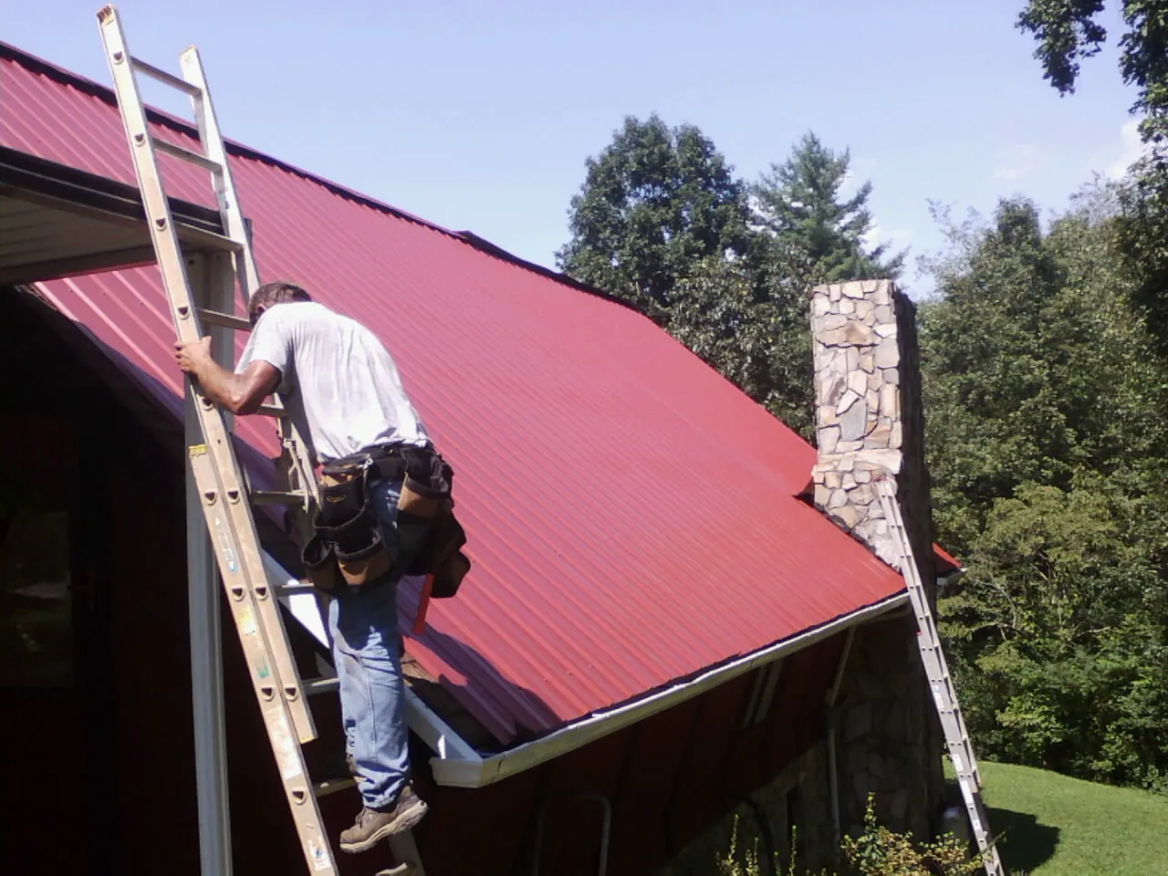 Roofer on a ladder beside a red metal roof and stone chimney — Soffit & Fascia Repair by Roofing Brothers in Euclid